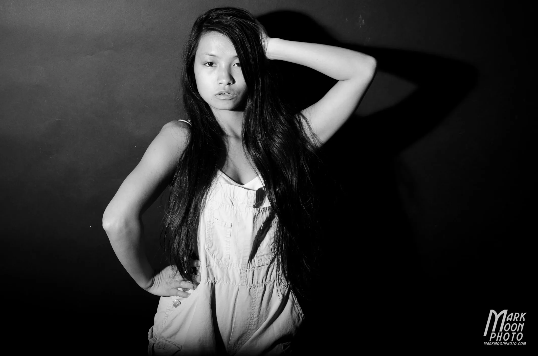 Black-and-white photo of Yi in overalls with a hand behind her head against a dark wall