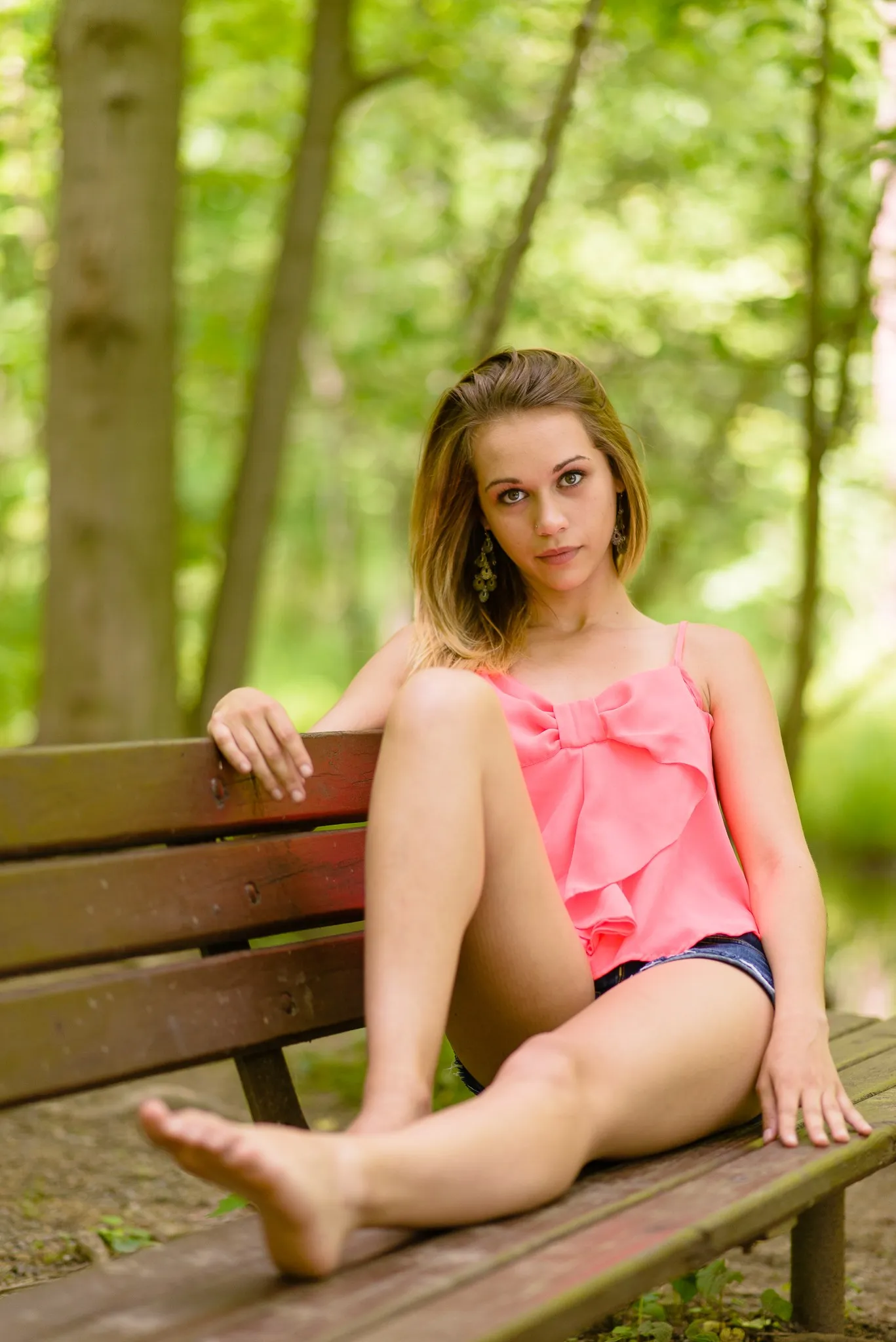 Aliyah sitting barefoot on a wooden park bench in a coral bow-front top and denim shorts, woods behind her
