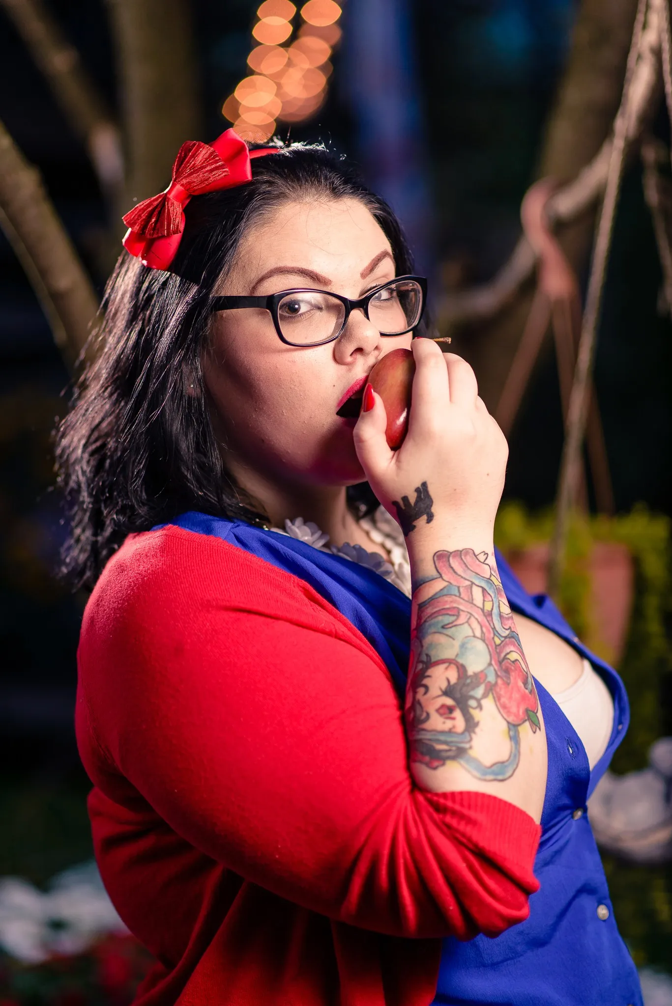 Carly biting a red apple, her tattooed forearm in front of her, string lights behind her