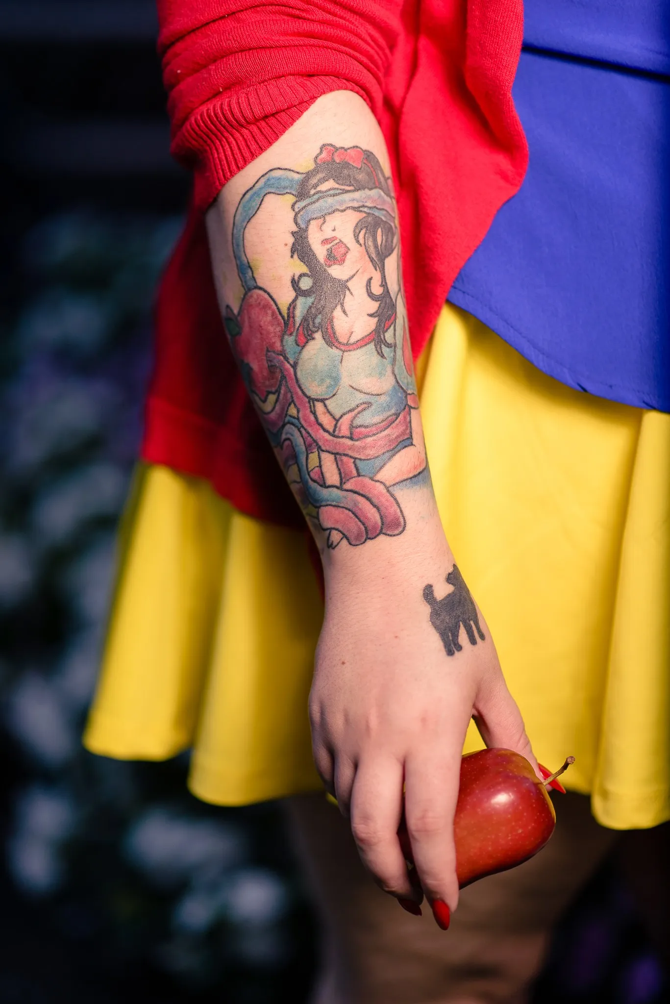 Close-up of the tattoo on Carly’s forearm, a red apple in her hand against her yellow skirt