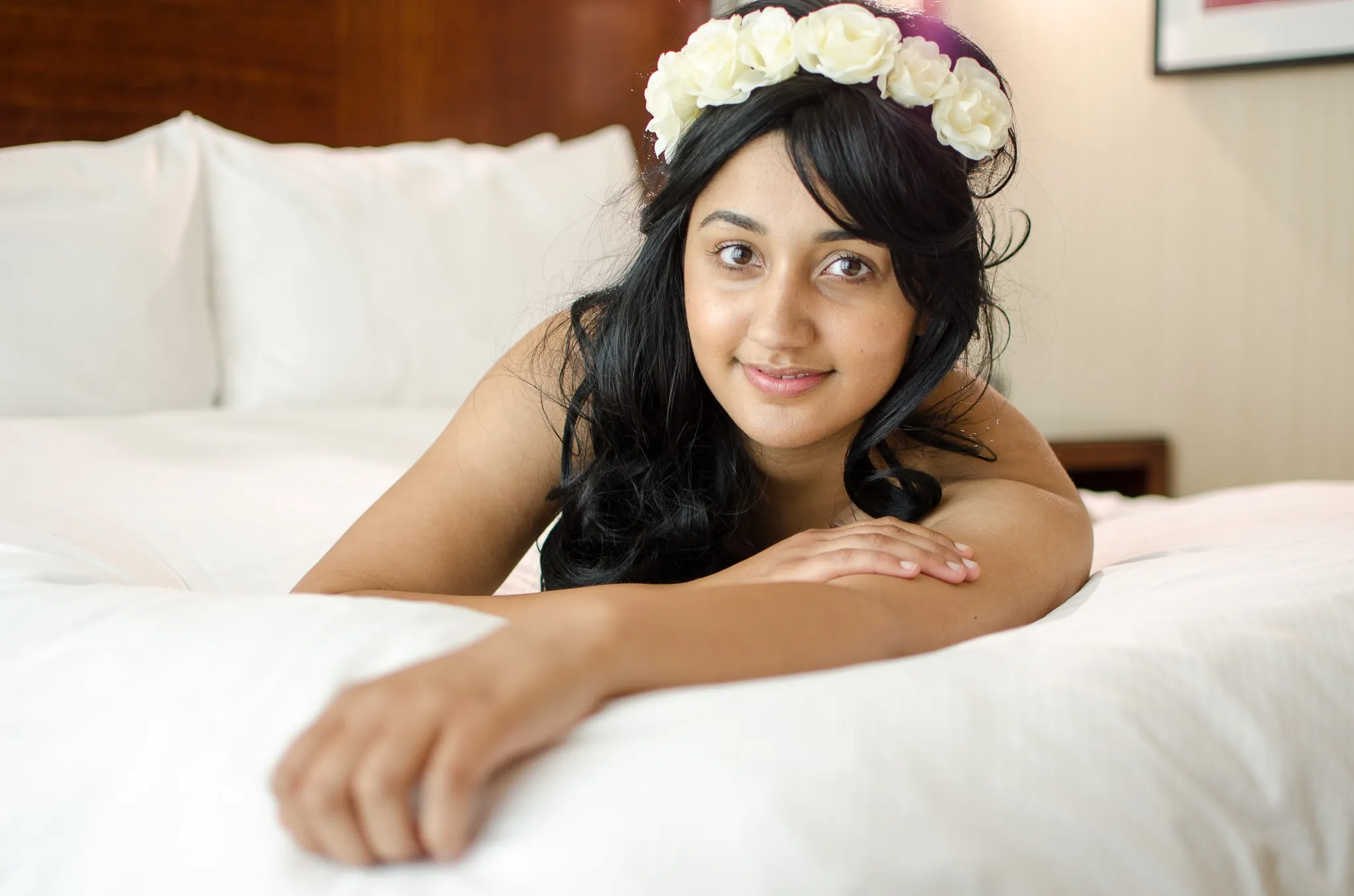 Elisha smiling, propped on her arms on a white bed, wearing a crown of white roses
