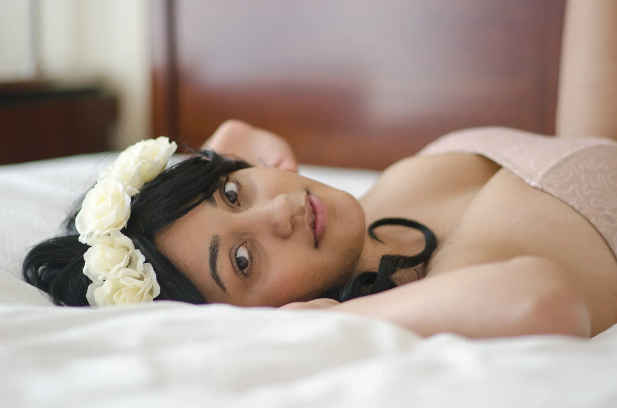 Elisha resting her head on a white bed, a crown of white roses in her dark hair