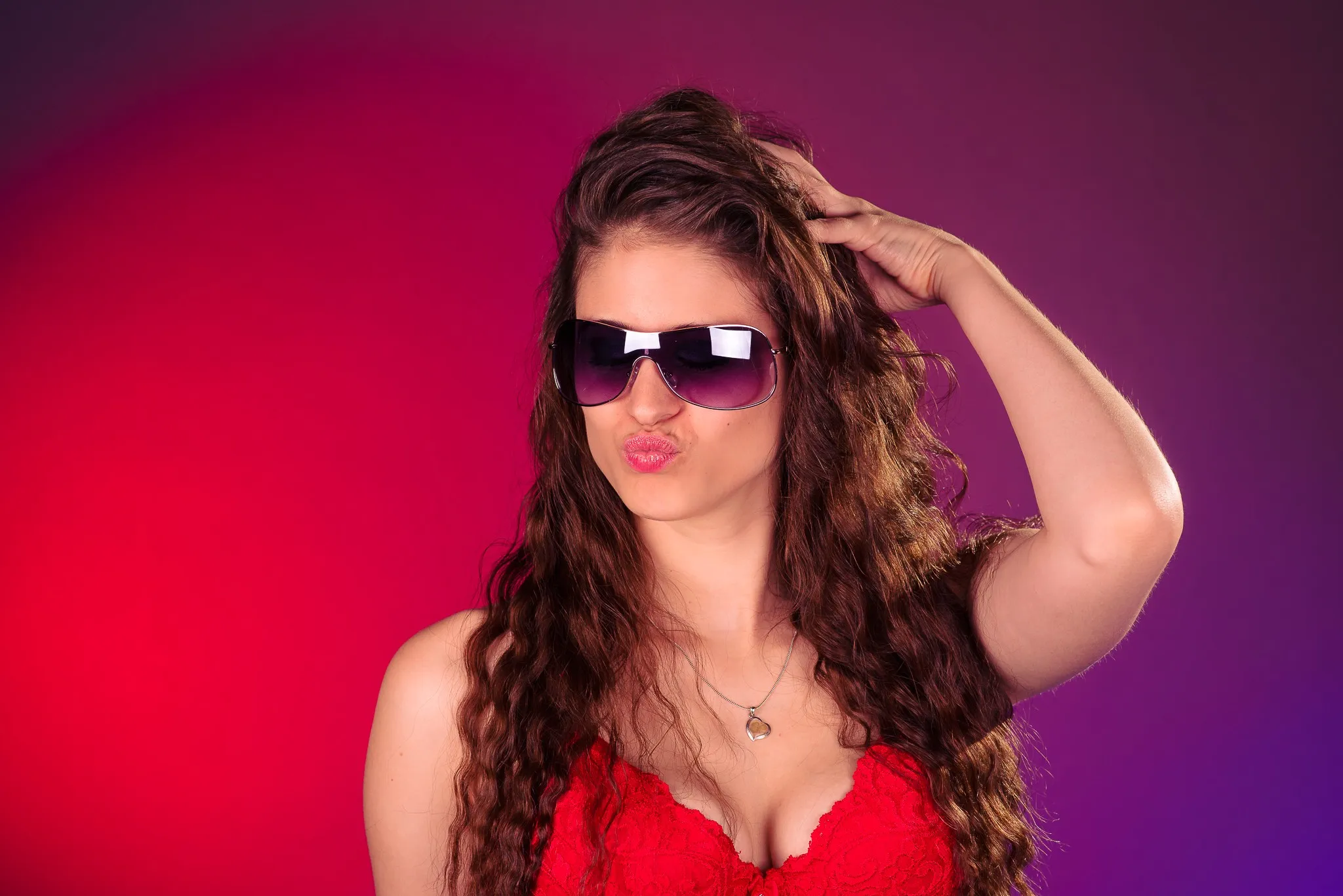 Kat pouting in sunglasses and a red lace bra, a hand in her hair, against a red and violet backdrop