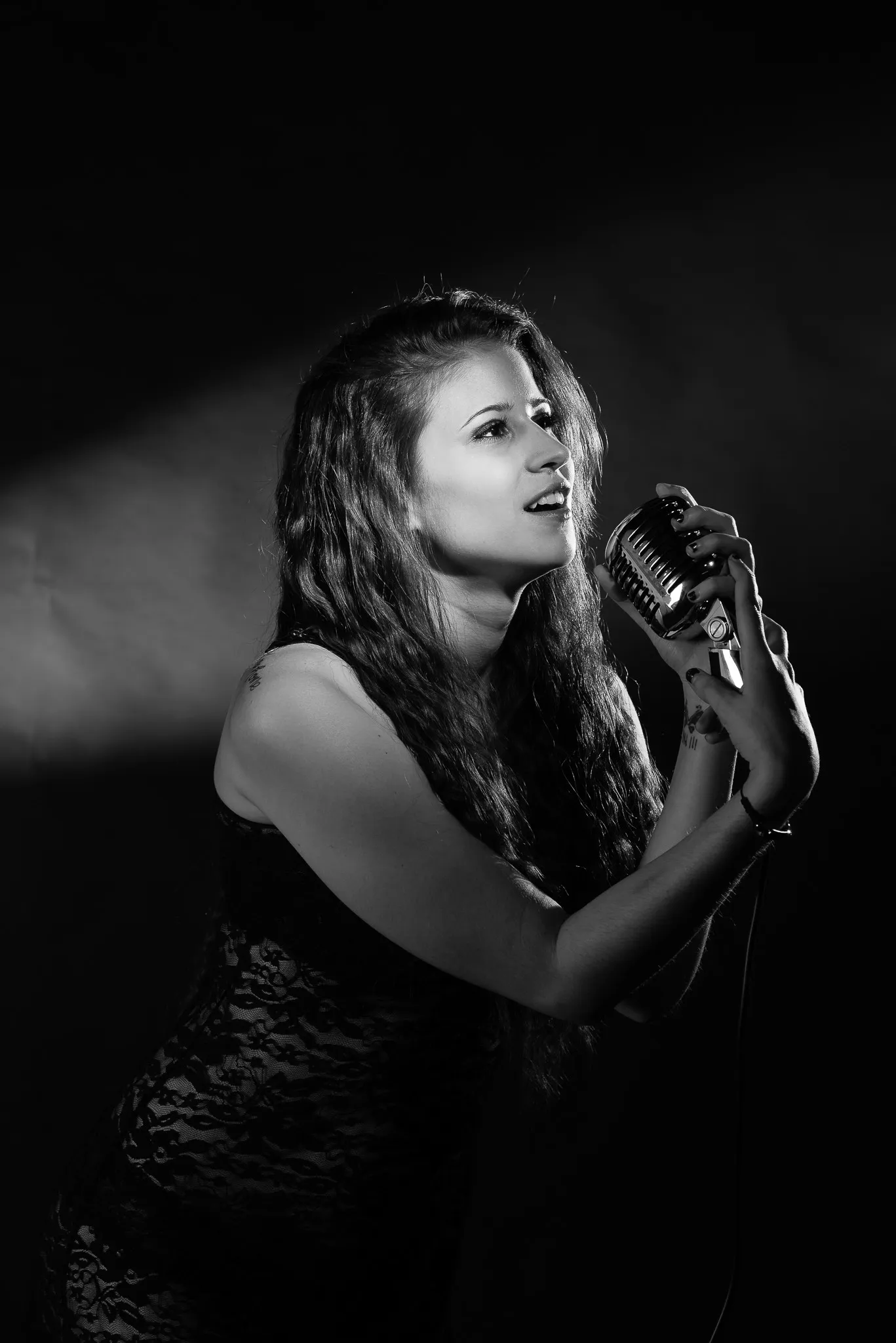 Black and white: Kat singing into a vintage microphone, a beam of light on the wall behind her