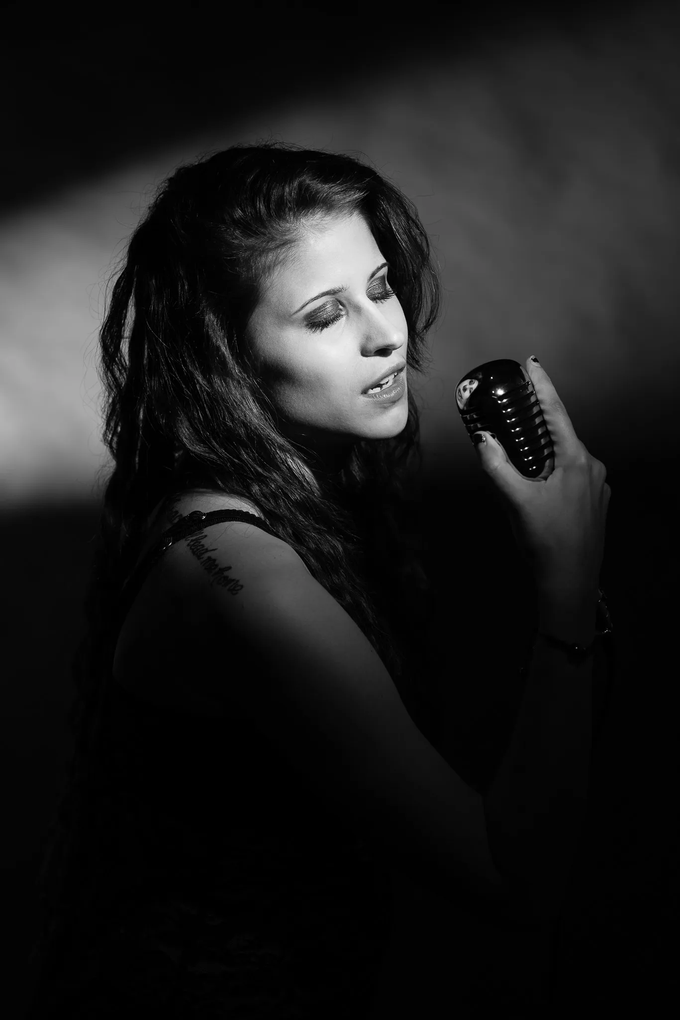 Black and white: Kat with her eyes closed, singing into a vintage microphone in a shaft of light