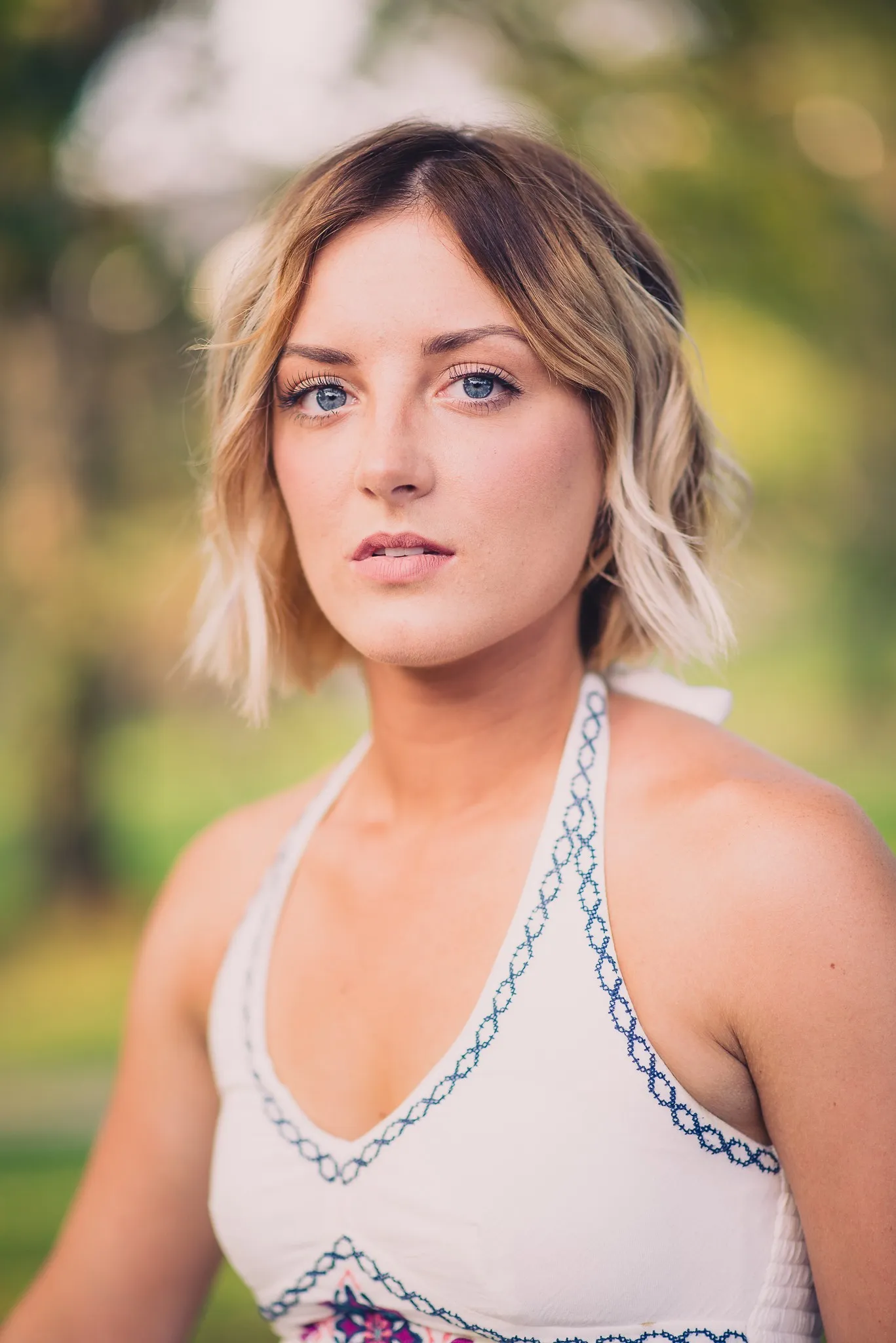 Close portrait of Sam with a blonde bob in a white embroidered halter dress, green trees behind her