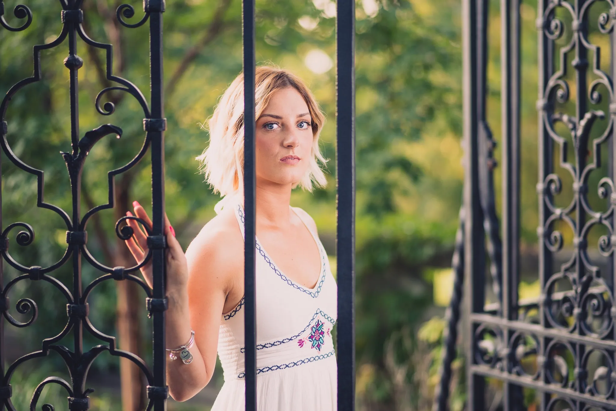 Sam in a white dress standing behind a wrought-iron gate in the park