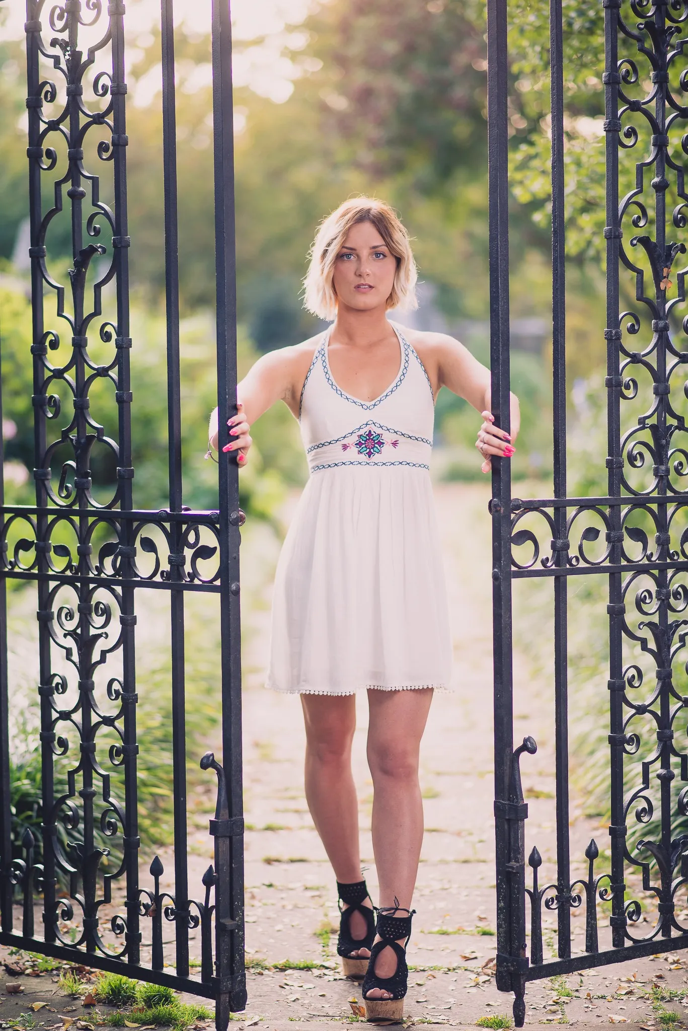 Sam in a white dress and black heels standing in an open wrought-iron gateway