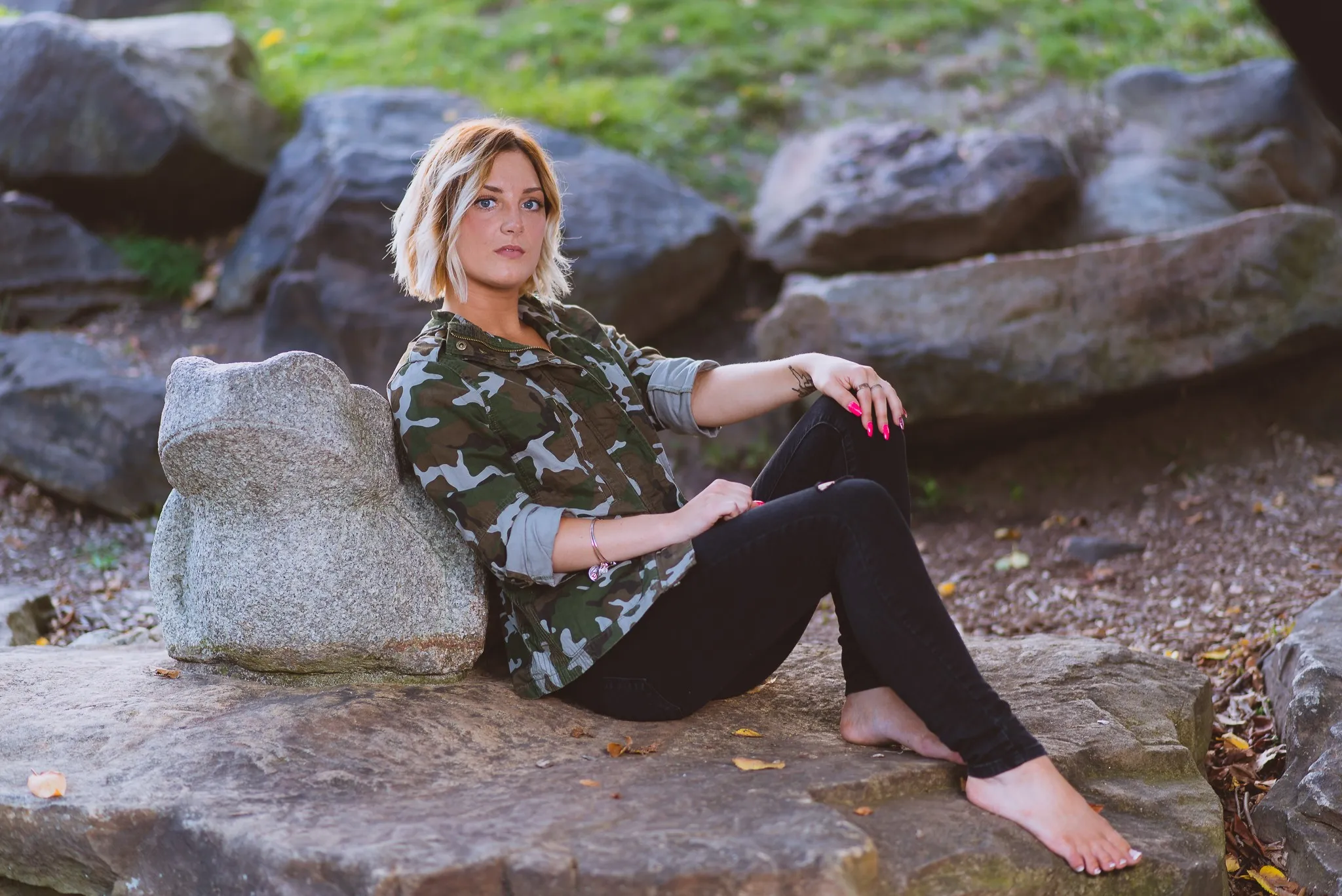 Sam in a camouflage jacket and black jeans sitting barefoot on boulders beside a stone sculpture