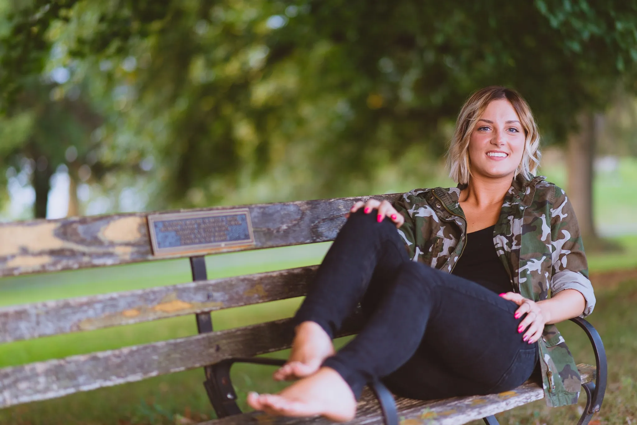 Sam laughing on a weathered park bench in a camouflage jacket, her bare feet up on the seat