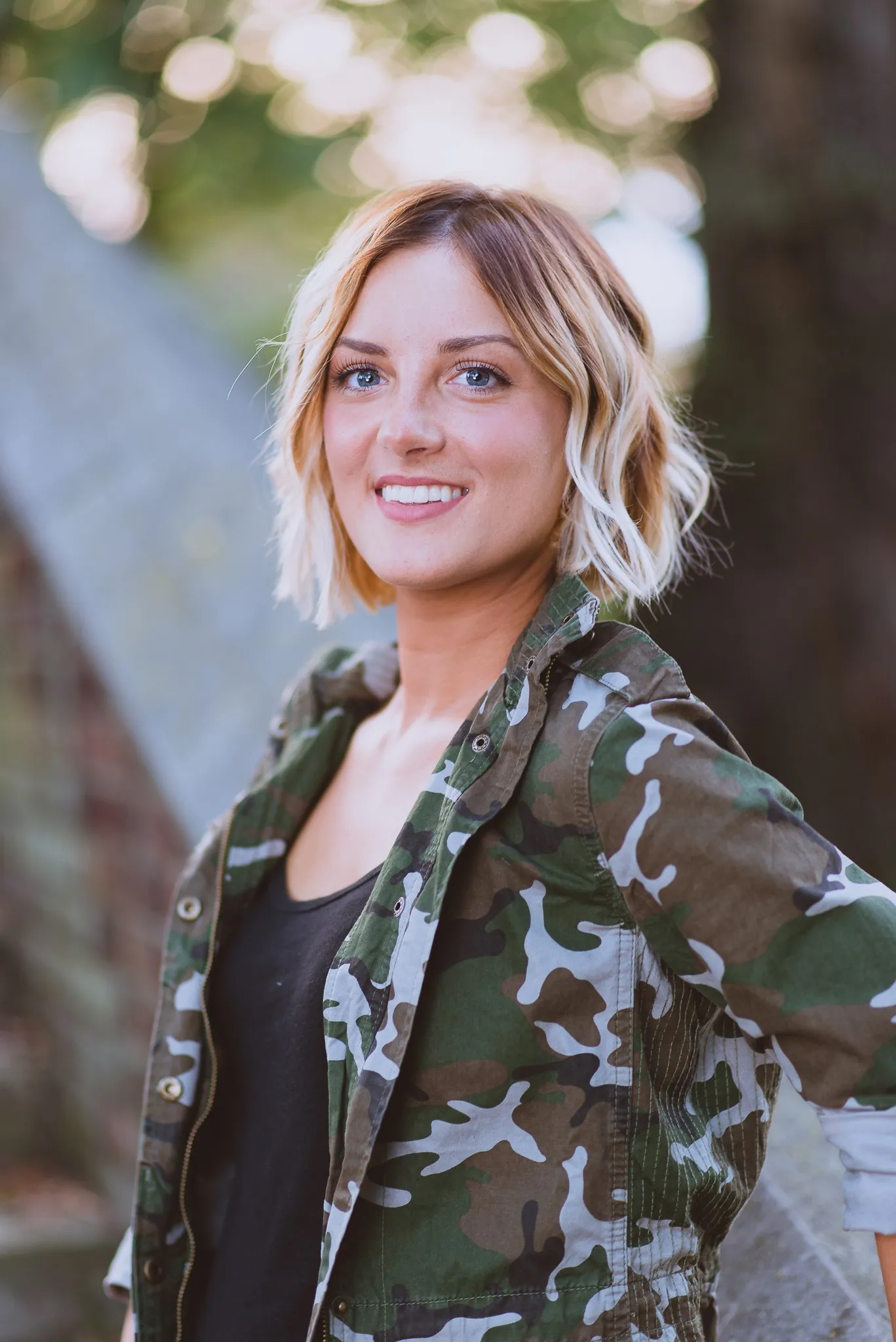 Sam smiling in a camouflage jacket beside a stone wall, sunlight in the trees behind her