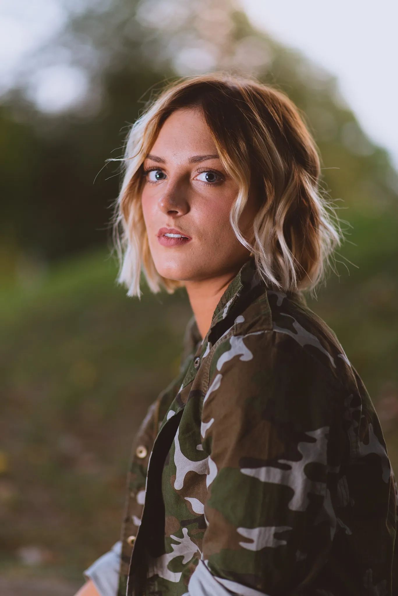 Sam looking back over her shoulder in a camouflage jacket at dusk